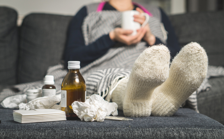 seorang wanita yang sakit terbaring di tempat tidur dengan kaus kaki hangat dan pil serta sirup di sampingnya