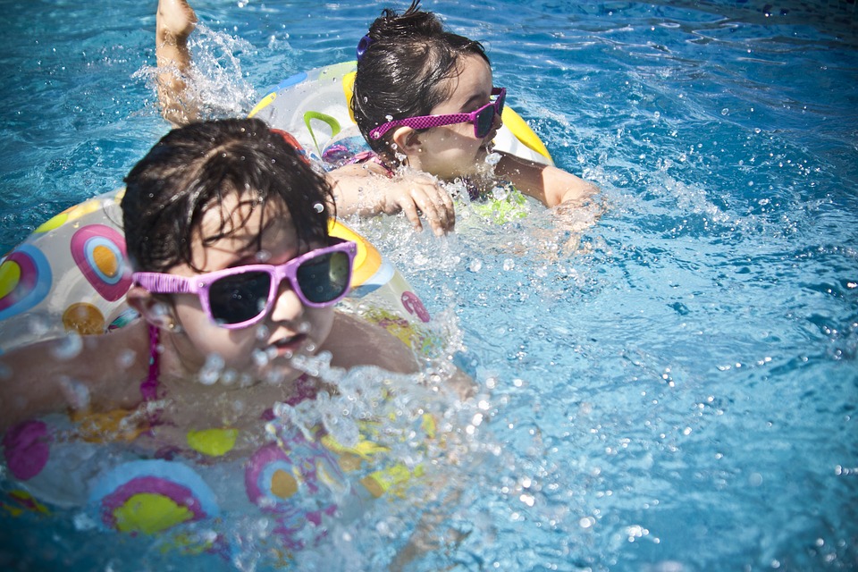 Dua orang anak sedang berenang di air, mereka mengenakan kacamata hitam dengan bingkai merah muda, dan mengendarai sepeda terapung.