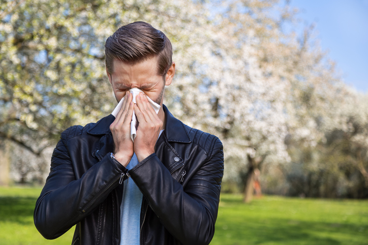 seorang pria dengan demam dan hidung penuh di dekat pohon yang sedang mekar dengan sapu tangan di tangannya