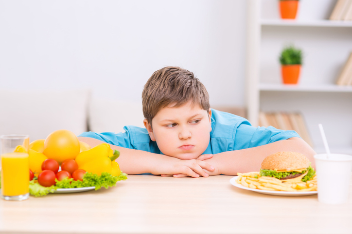 Seorang anak gemuk di antara dua piring makanan sehat dan tidak sehat.