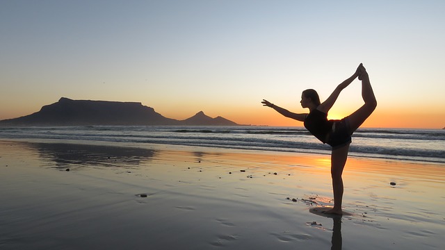 Seorang wanita sedang berlatih yoga di pantai. Matahari telah terbenam. Masih terang. Dia berdiri dengan satu kaki. Air laut sedang surut. Pasir berada di bawah lapisan air yang tipis.