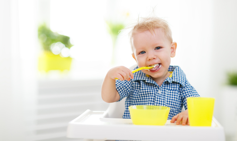 Seorang anak kecil yang duduk di kursi bayi, sedang menyuapi dirinya sendiri. Sendok, cangkir, dan mangkuk berwarna kuning